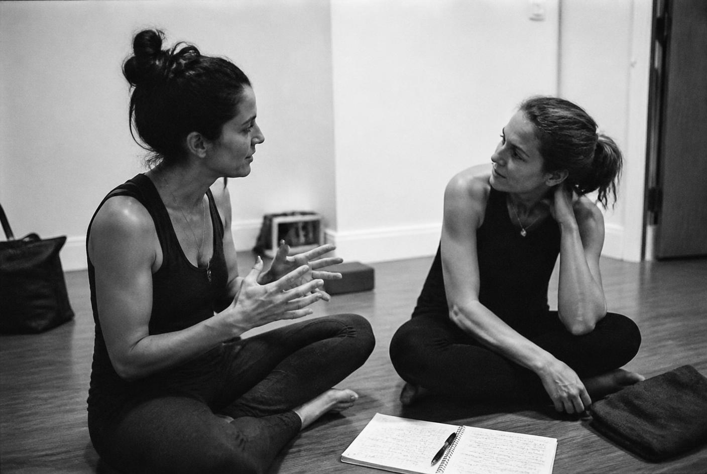 Two yoga teachers sitting on a studio floor in conversation
