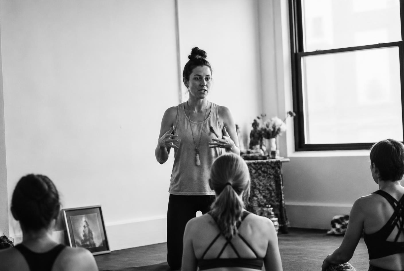 A yoga teacher speaking to a class from the front of a studio